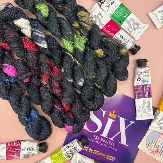 A styled flat-lay featuring several skeins of variegated yarn in dark charcoal, black, green, pink, and blue, arranged alongside tubes of acrylic and watercolor paints and a purple 'Six The Musical' poster on a light pink background.