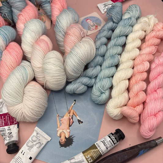 A flat lay of pastel yarn skeins in soft pink, blue, cream, and white, arranged with art supplies including paint tubes and a small printed card on a pink background.